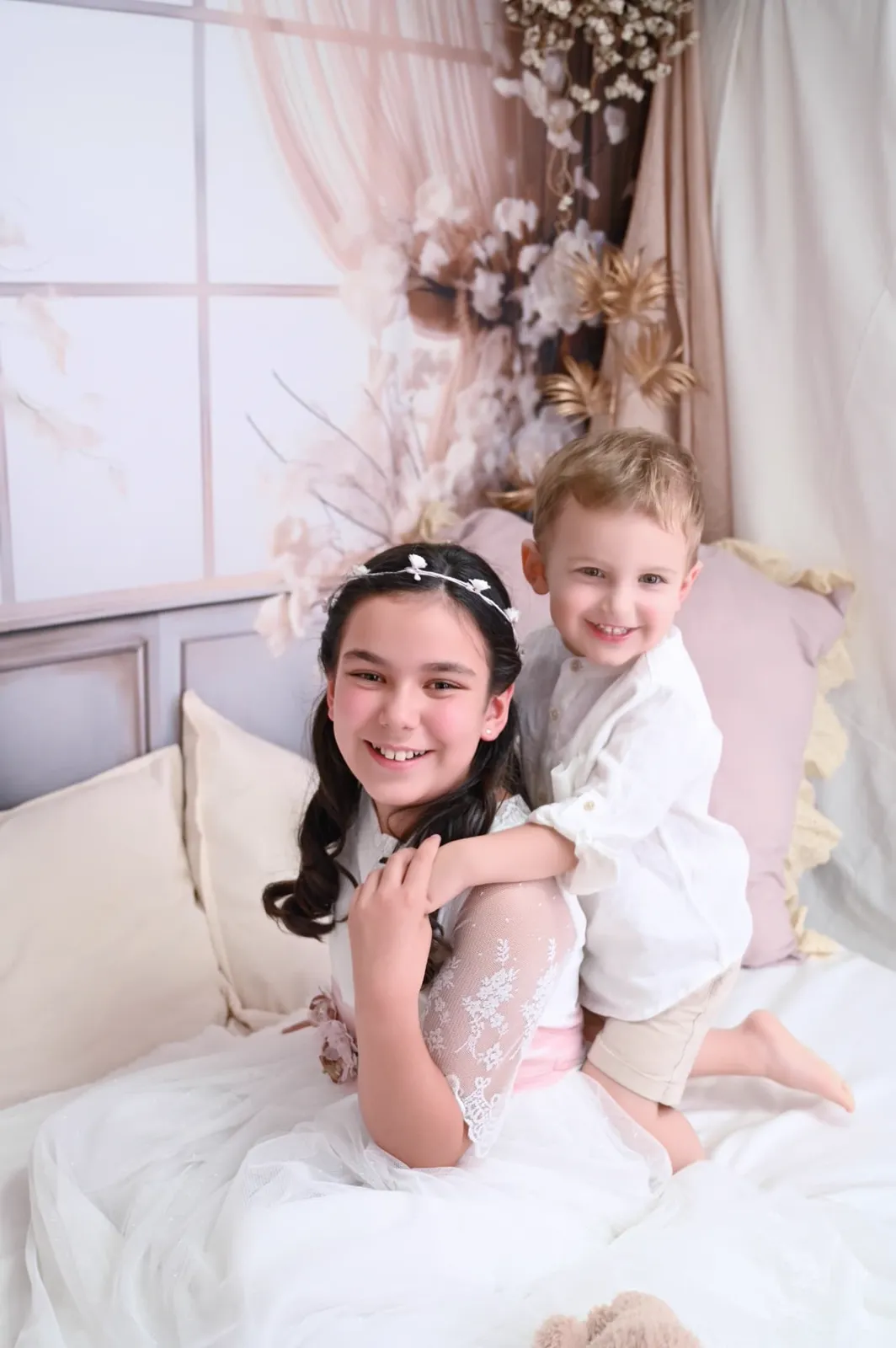 Niña de Comunión sentada en el set de camita del estudio con su hermano pequeño abrazándola