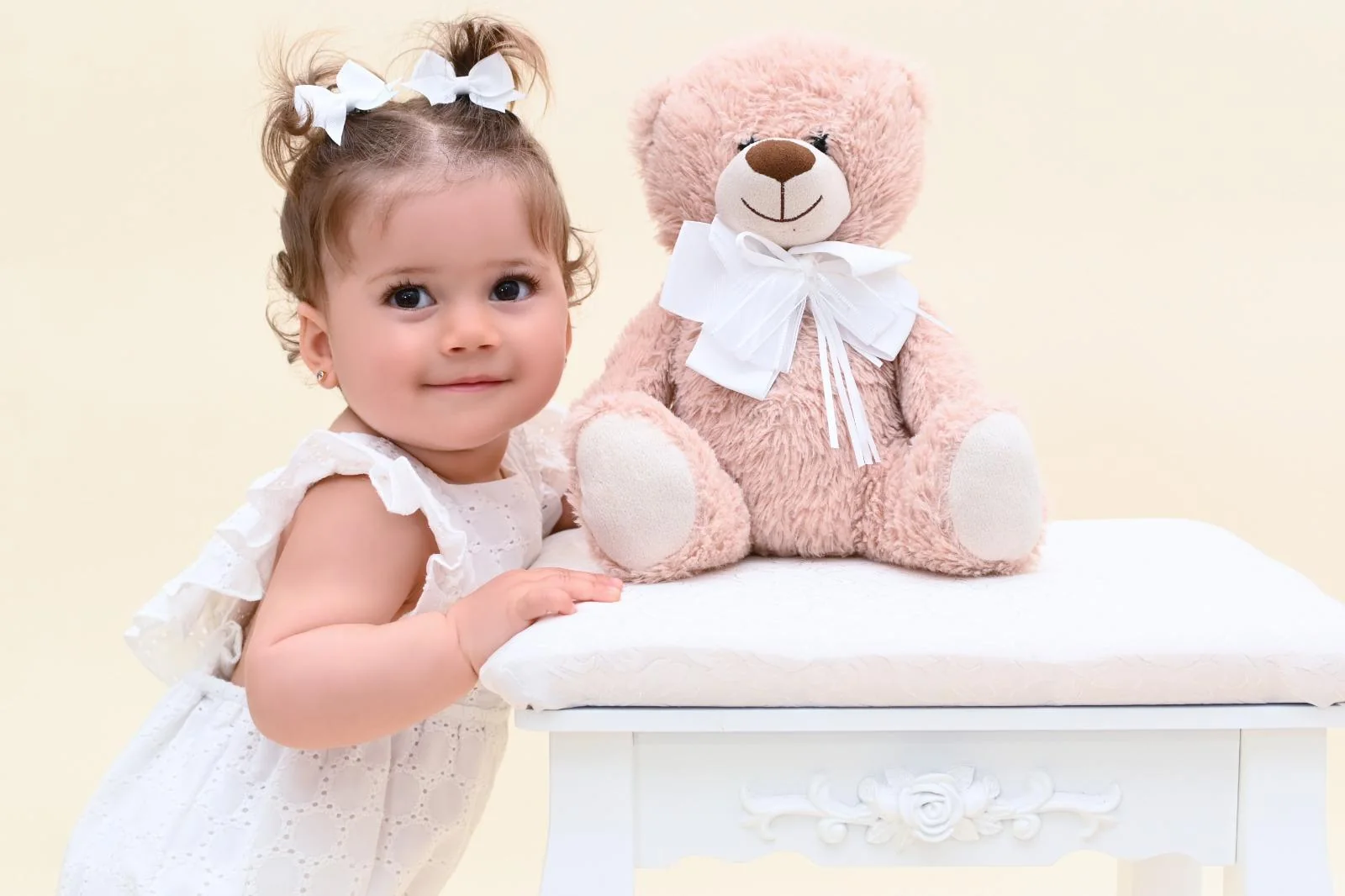 Niña de un año con vestido blanco junto a un osito de peluche en el estudio