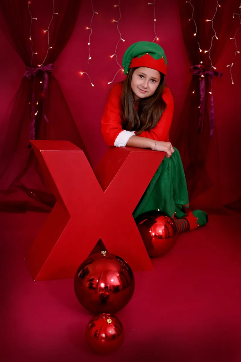 Niña vestida de elfa apoyada en una gran letra X roja en el decorado XMAS del estudio de Fuenlabrada