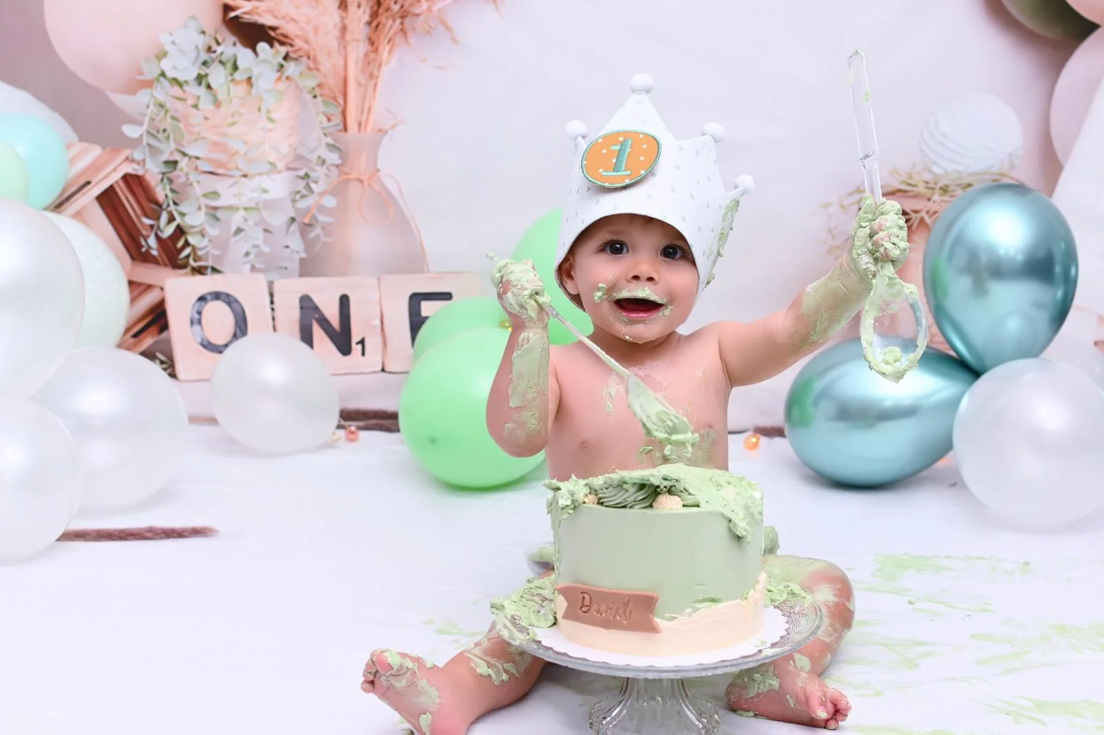 Bebé con las manos llenas de crema verde levantando dos cucharas durante su Smash Cake