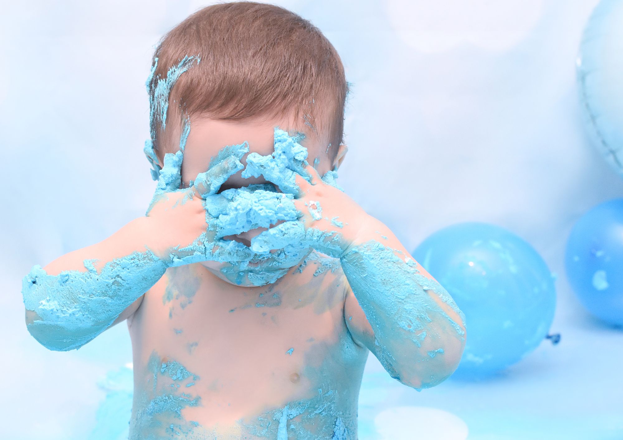 Bebé cubierto de crema azul tapándose la cara durante la sesión de smash cake