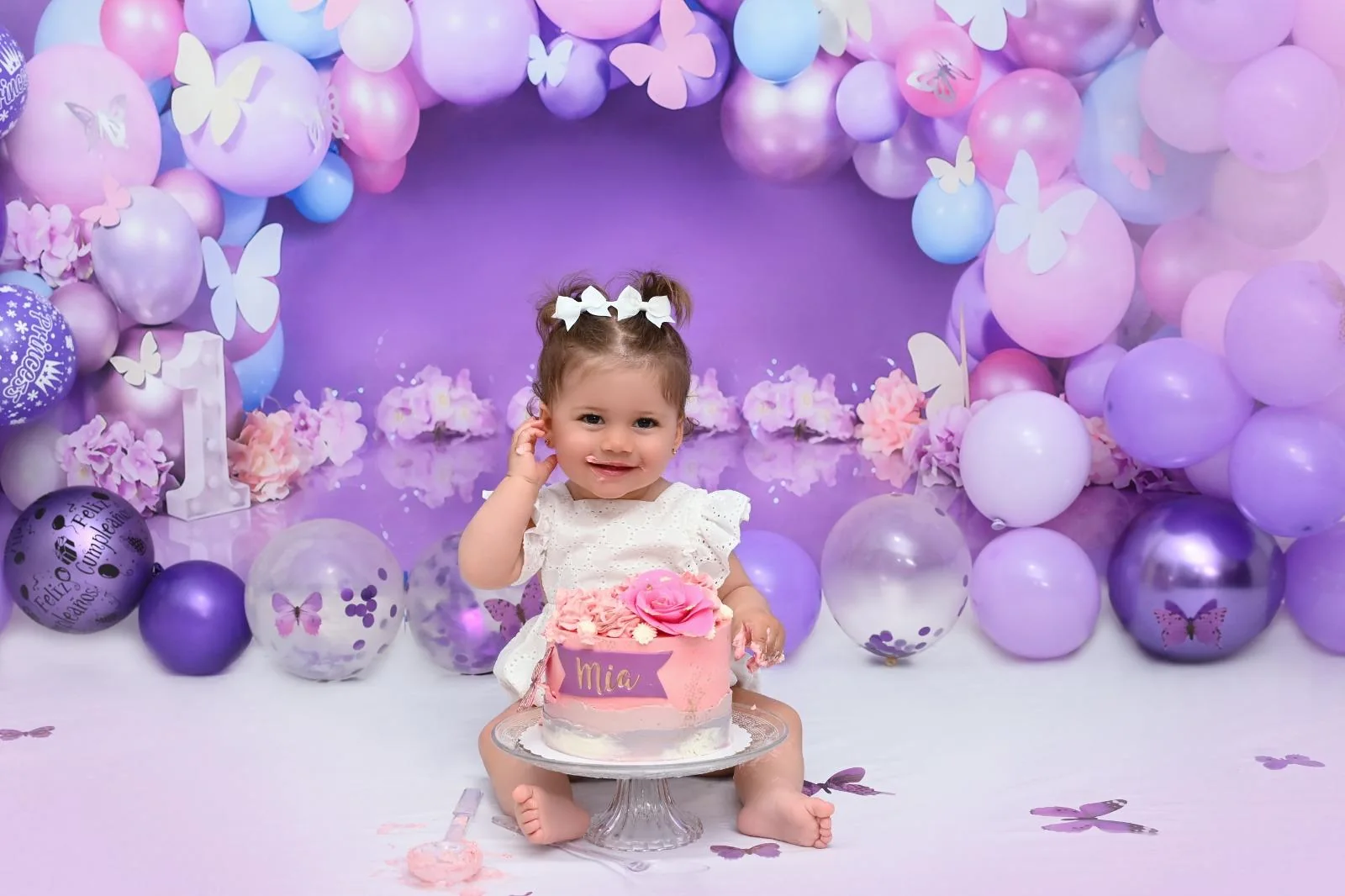 Bebé con lazos blancos celebrando su primer cumpleaños con tarta rosa en un decorado de mariposas