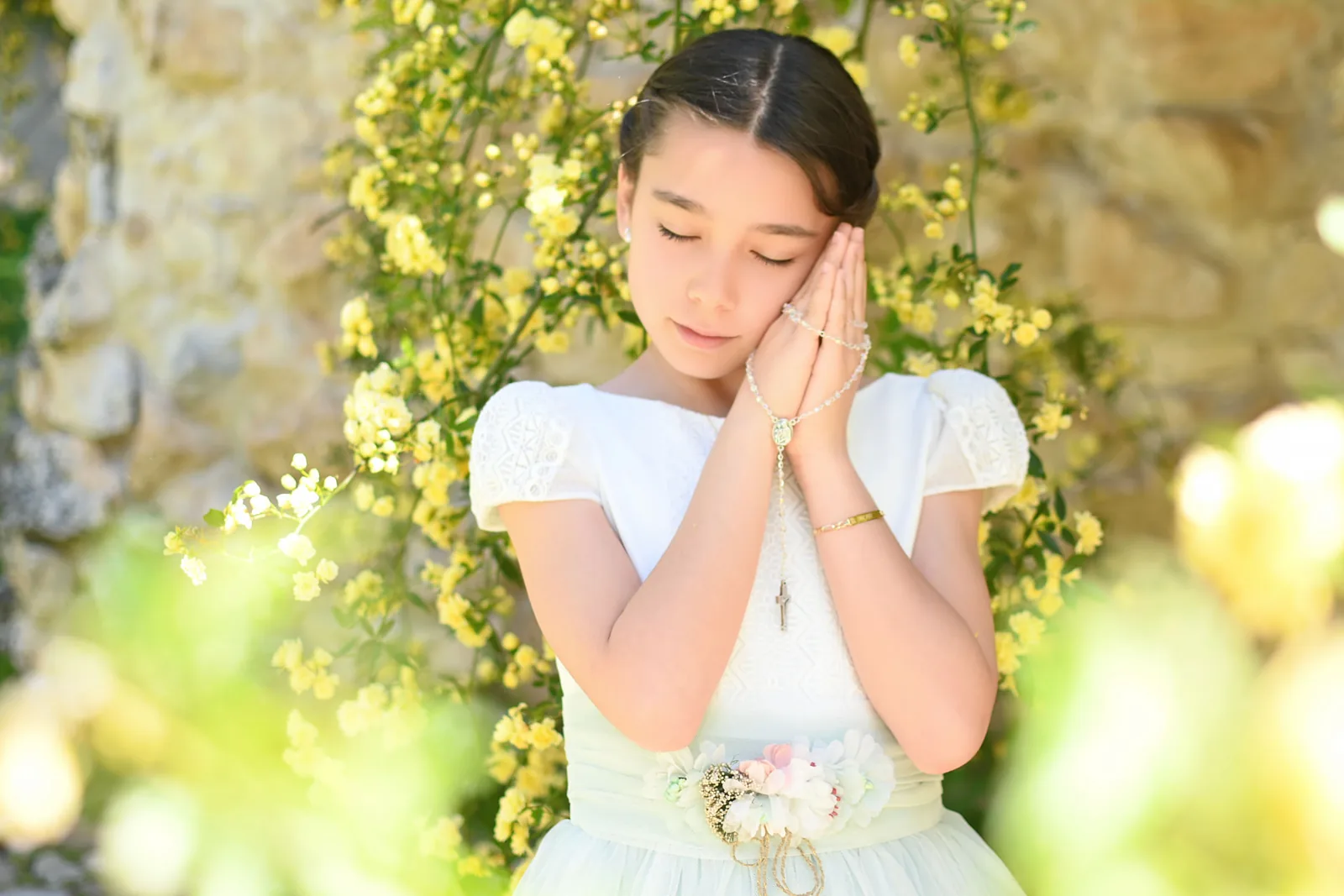 Niña de Primera Comunión con los ojos cerrados y las manos juntas entre flores amarillas
