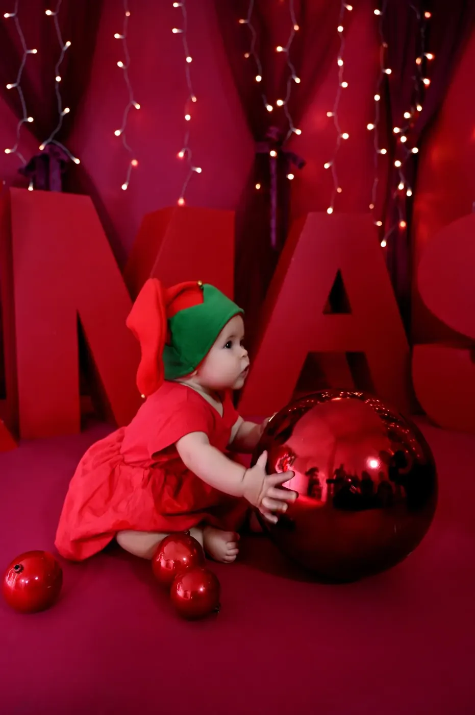 Bebé vestido de elfo junto a una gran bola roja de Navidad
