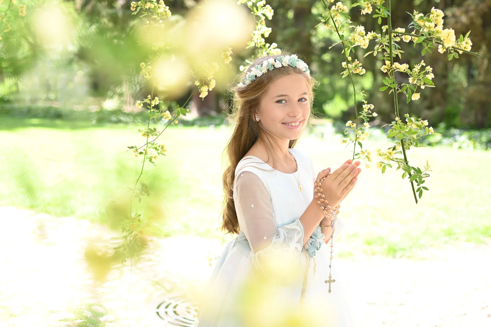 Niña de Comunión sonriendo con un rosario entre las manos bajo ramas de flores amarillas