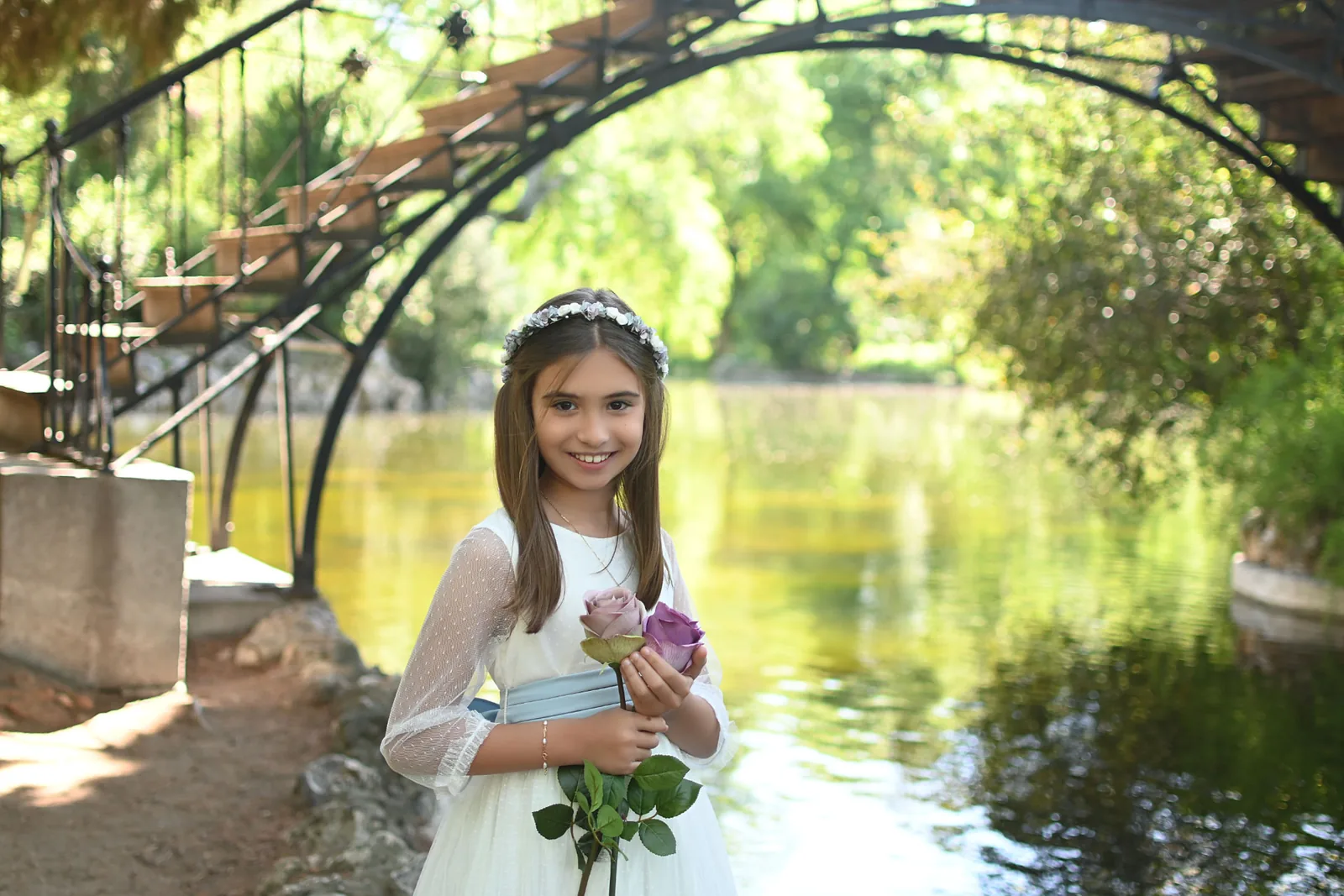 Niña de Primera Comunión con rosas junto a un lago y un puente de hierro durante una sesión en exterior