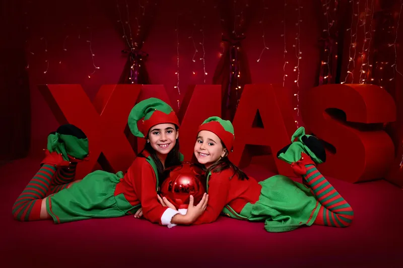 Dos hermanas vestidas de elfas tumbadas abrazando una bola roja delante de las letras XMAS en Fuenlabrada