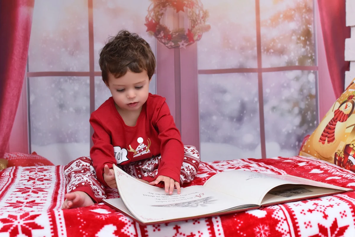Niño con pijama rojo leyendo un libro sobre una cama con decoración navideña, campaña 2020
