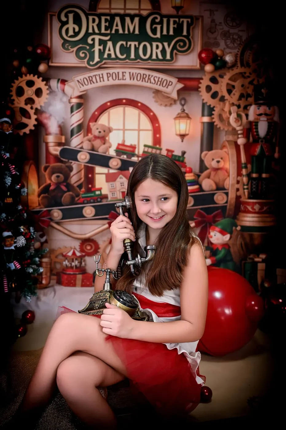 Niña con vestido rojo y blanco sonriendo con un teléfono antiguo en el decorado de la fábrica de juguetes de la Navidad 2026