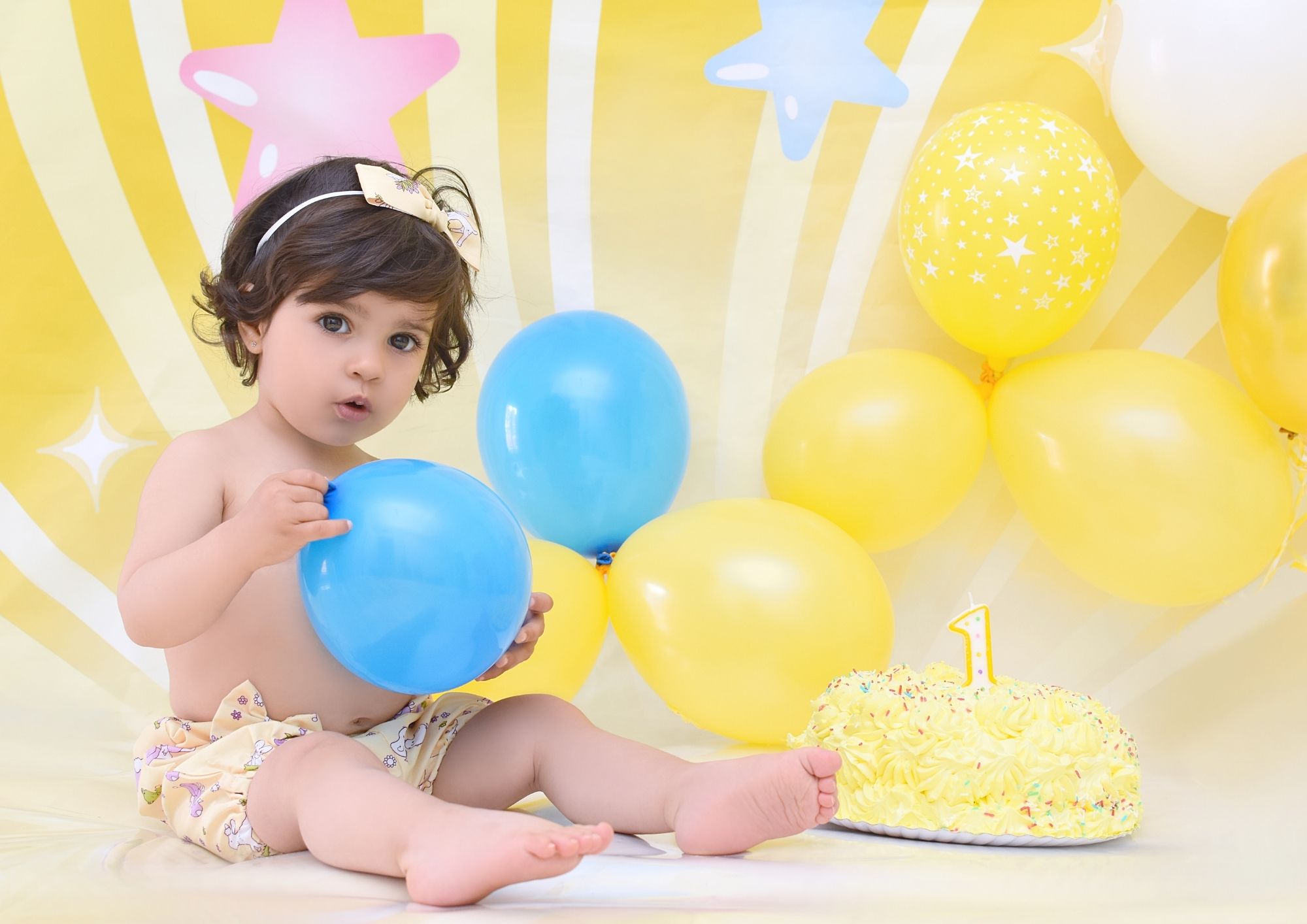 Bebé con globo azul junto a su tarta de primer cumpleaños en un decorado amarillo de estrellas, sesión Smash Cake en Fuenlabrada
