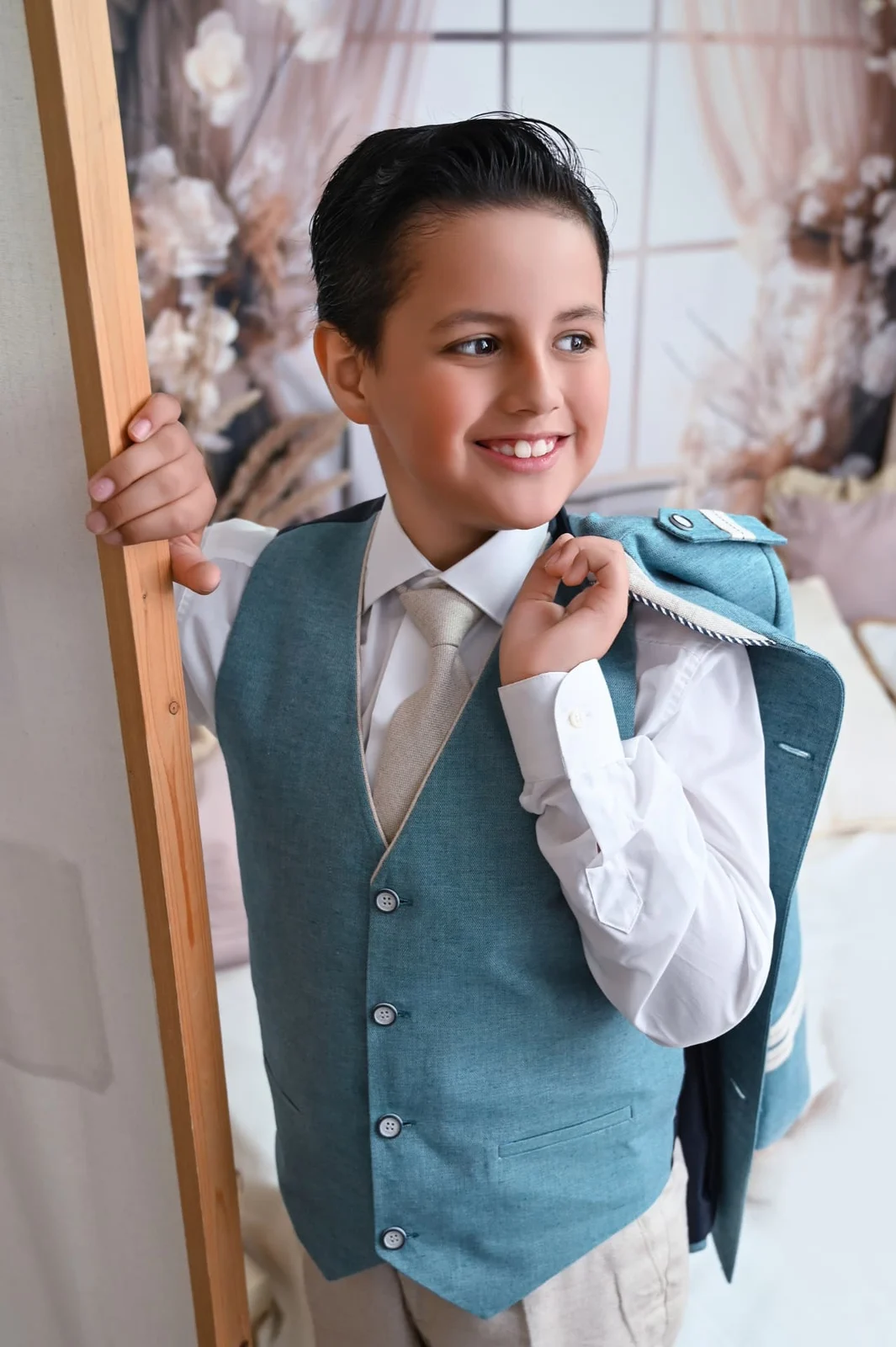 Niño de Comunión sonriendo con chaleco azul y la chaqueta al hombro en un set floral del estudio